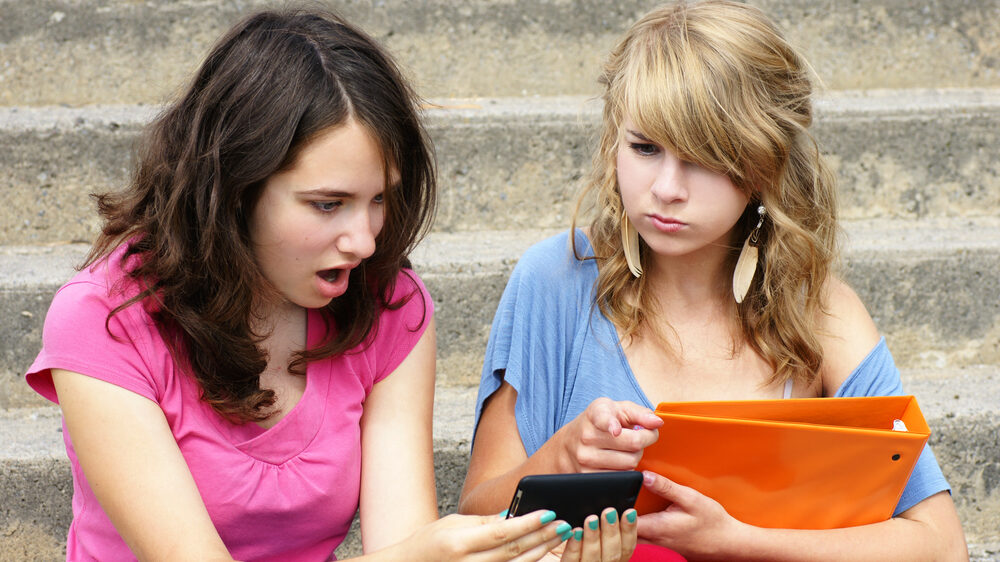 Image of two teen girls sitting on steps, looking at a cell phone....