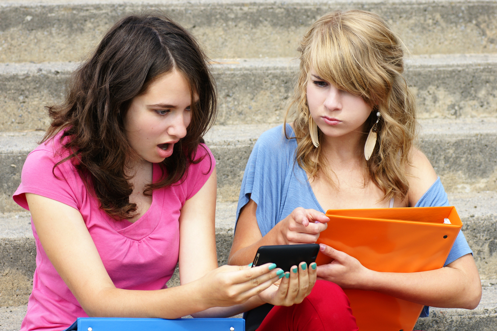 Cyber or online bullying concept with two young women students or teenager girls shocked at the text they are reading on their cell phone, perfect for awareness.