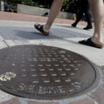 Pedestrians walk past a manhole cover for a sewer in Berkeley, Calif., Thursday, July 18, 2019. Soon students in Berkeley, California will have to pledge to "collegiate Greek system residences" instead of sororities or fraternities and city workers will have to refer to manholes as "maintenance holes." Officials in the liberal city this week passed an ordinance to replace some terms with gender-neutral words in the city code. (AP Photo/Jeff Chiu)
