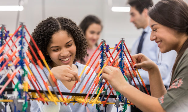 Young women enjoy building a model of a bridge during engineering class. They are smiling cheerfull...