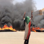FILE - In this June 3, 2019 file photo, a protester wearing a Sudanese flag flashes the victory sign in front of burning tires and debris, near Khartoum's army headquarters, in Khartoum, Sudan. Sudan’s uprising has ushered in a new era both for the nation and for Sudanese women after three decades of autocratic rule by Omar al-Bashir. Sudanese women played a pivotal role in the protests that brought down al-Bashir, and under a joint military-civilian council in power now, they hope for more freedom and equality, and seek to overturn many of the restrictive Islamic laws from the previous era. (AP Photo, File)