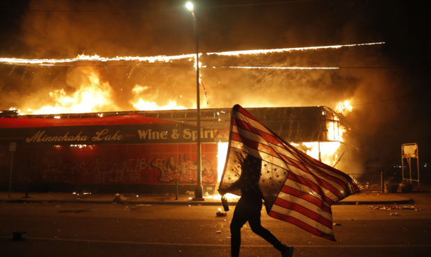 A protester carries the carries a U.S. flag upside, a sign of distress, next to a burning building ...