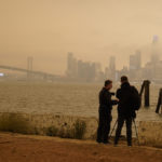 Two men adjust a camera while shooting pictures of wildfire smoke over the darkened skyline from Treasure Island Wednesday, Sept. 9, 2020, in San Francisco. People from San Francisco to Seattle woke Wednesday to hazy clouds of smoke lingering in the air, darkening the sky to an eerie orange glow that kept street lights illuminated into midday, all thanks to dozens of wildfires throughout the West. (AP Photo/Eric Risberg)