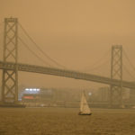 Under darkened skies from wildfire smoke, a sailboat makes its way past the San Francisco-Oakland Bay Bridge and lights at Oracle Park Wednesday, Sept. 9, 2020, in San Francisco. People from San Francisco to Seattle woke Wednesday to hazy clouds of smoke lingering in the air, darkening the sky to an eerie orange glow that kept street lights illuminated into midday, all thanks to dozens of wildfires throughout the West. (AP Photo/Eric Risberg)