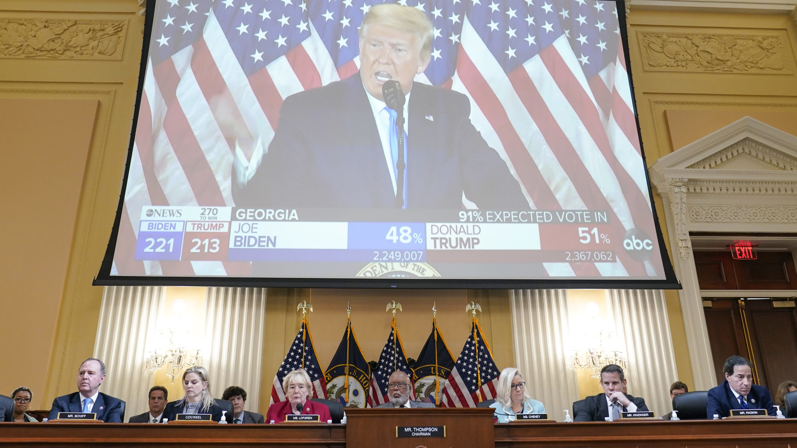 A large screen showing President Trump sits above the committee members presenting the Jan. 6 heari...