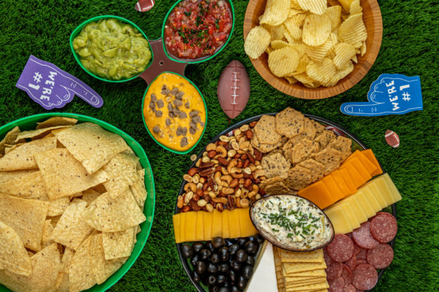 Display of bowls of chips, platter of cheese, crackers, olives, nuts, salami and nuts with spinach articoke dip, salsa, cheese dip and guacamole and football tailgate themed party decorations horizontal orientation