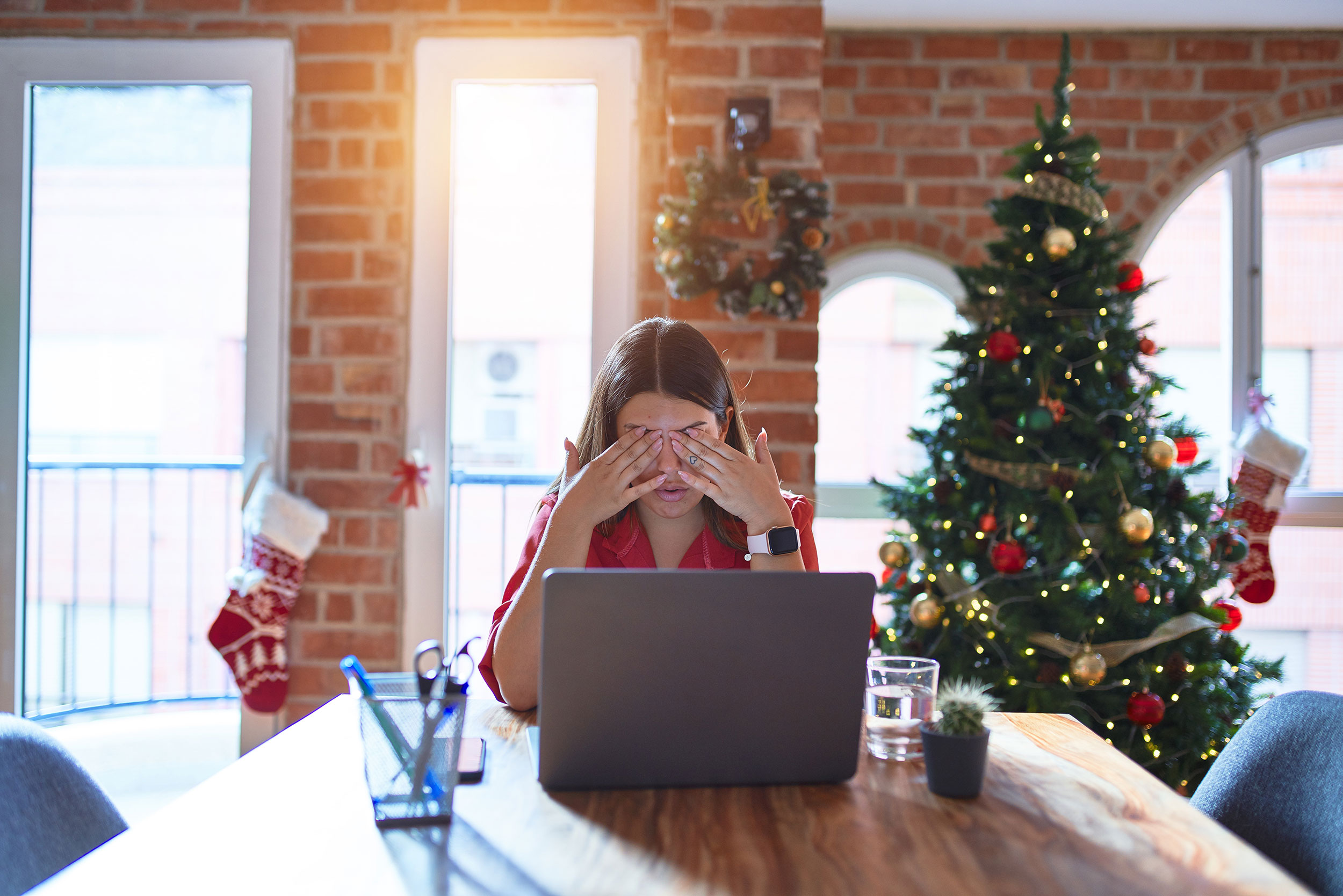 Beautiful woman sitting at the table working with laptop at home around christmas tree rubbing eyes...