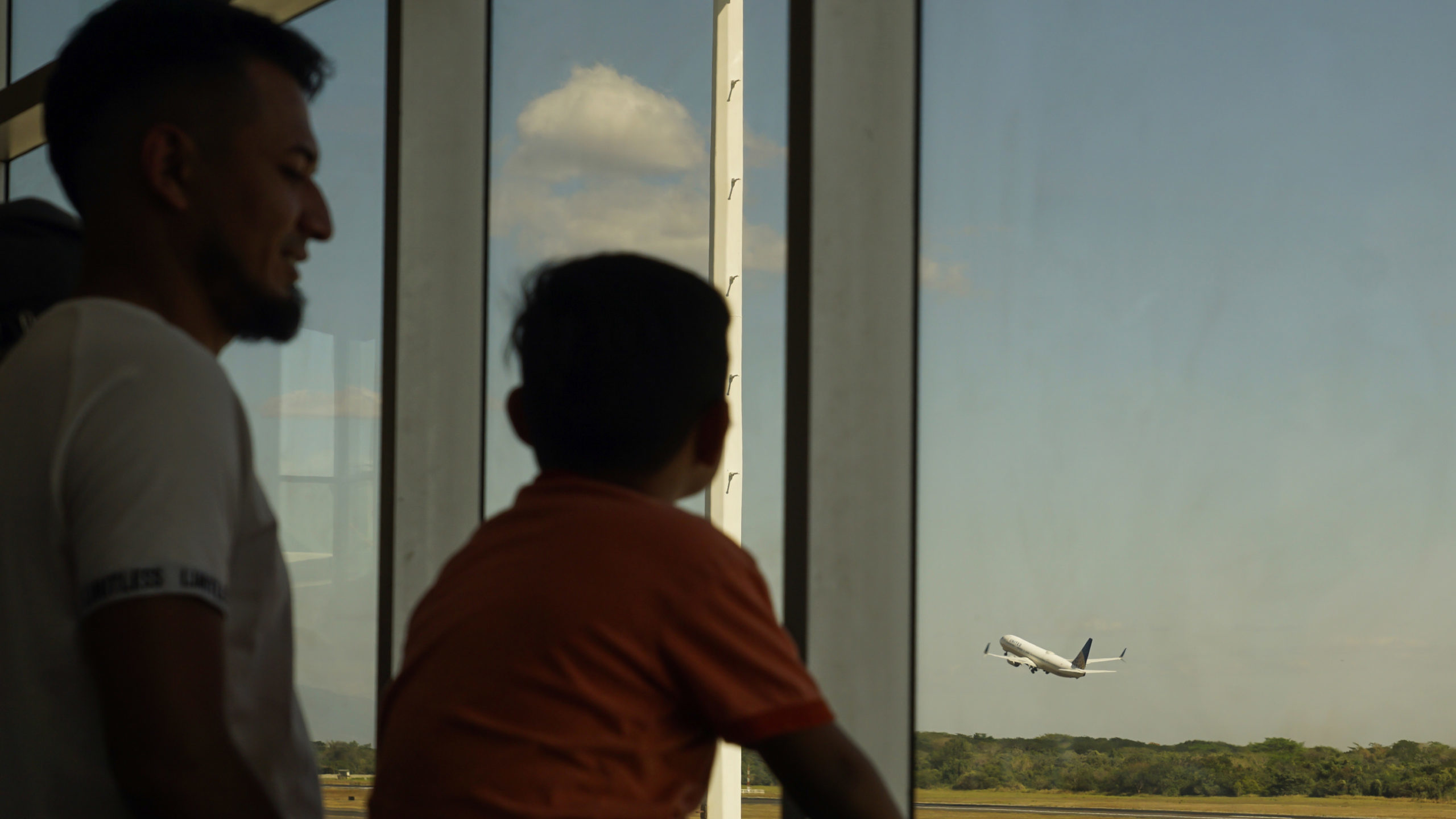 A man and his son watch a United Airlines plane take off from Comalapa International Airport on Feb...