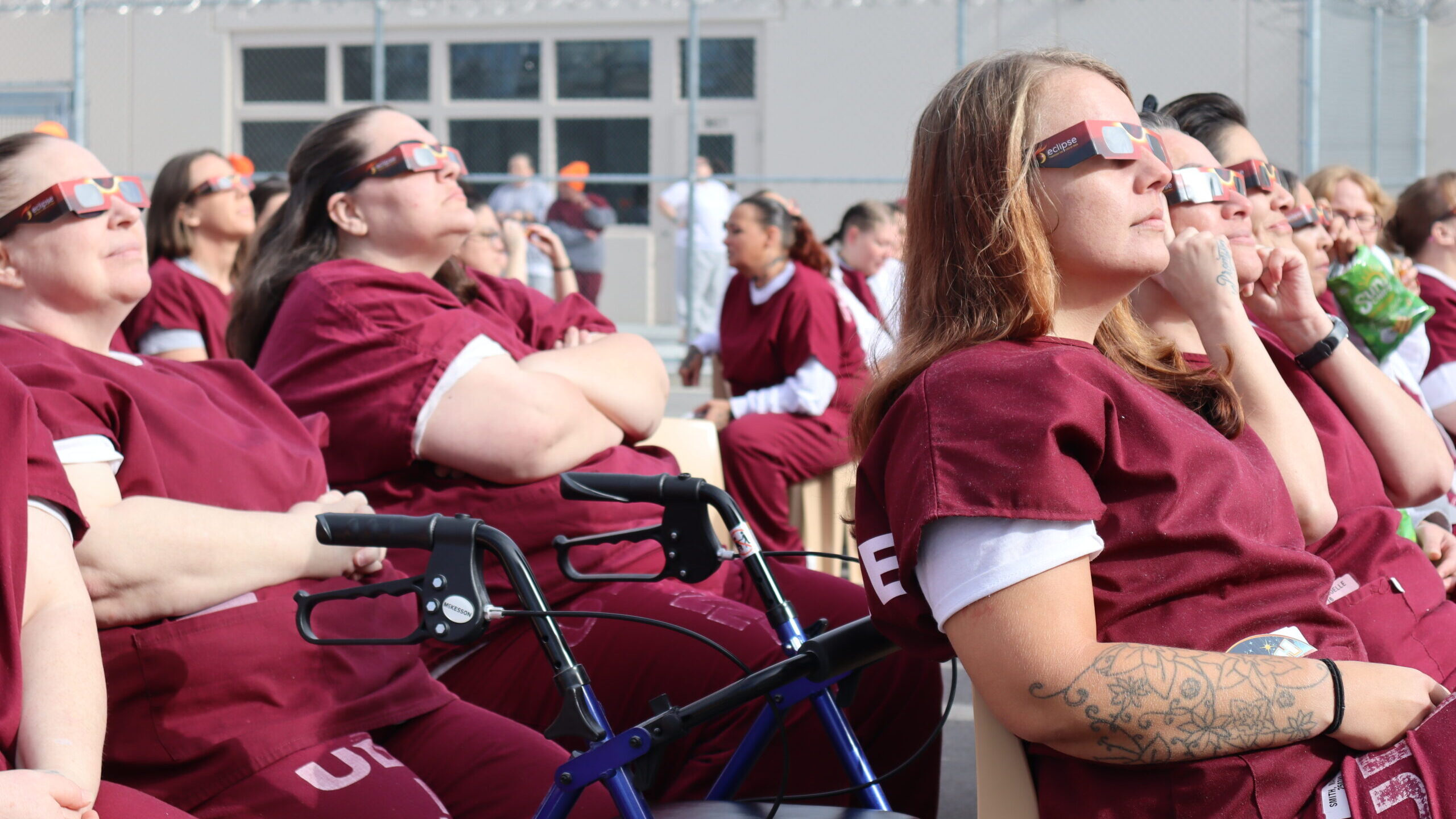 Image of several incarcerated women sitting outside in their prison jumpsuits and protective glasse...