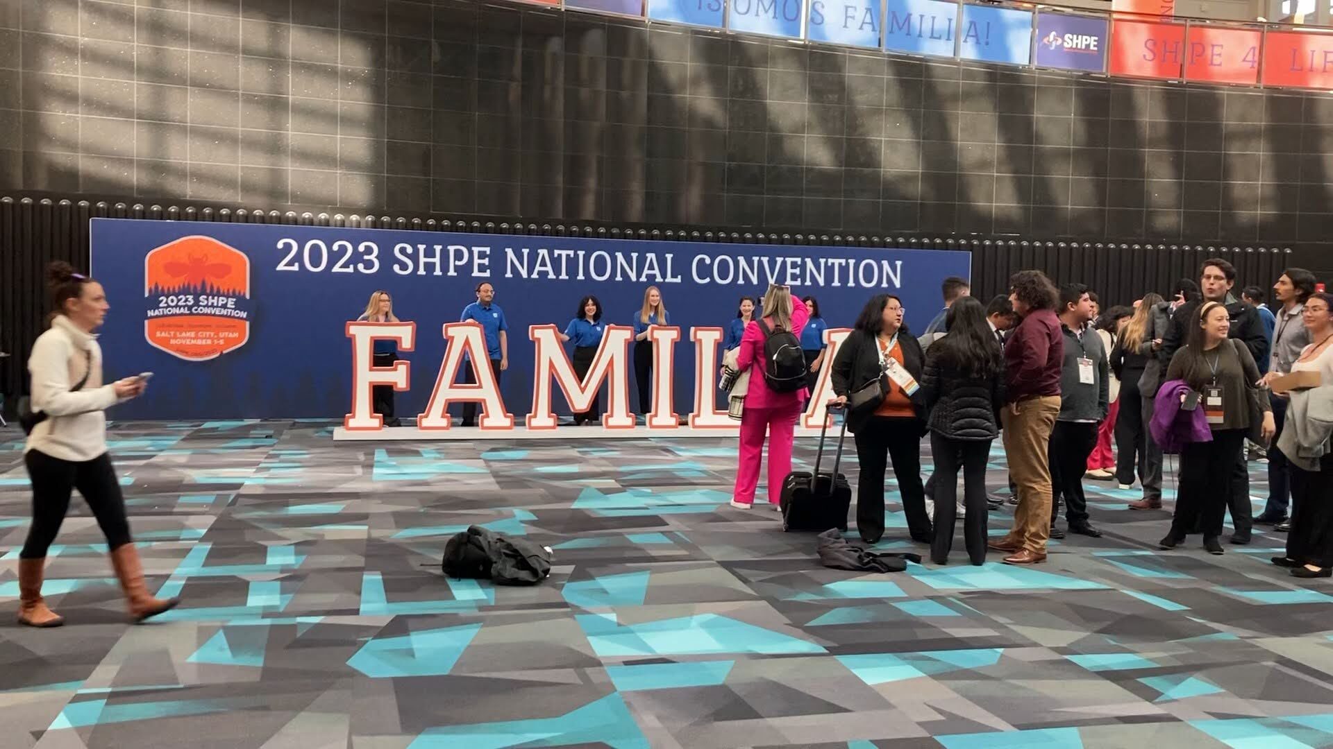 several people walk past a big sign that says "familia," as they attend the SHPE conference....