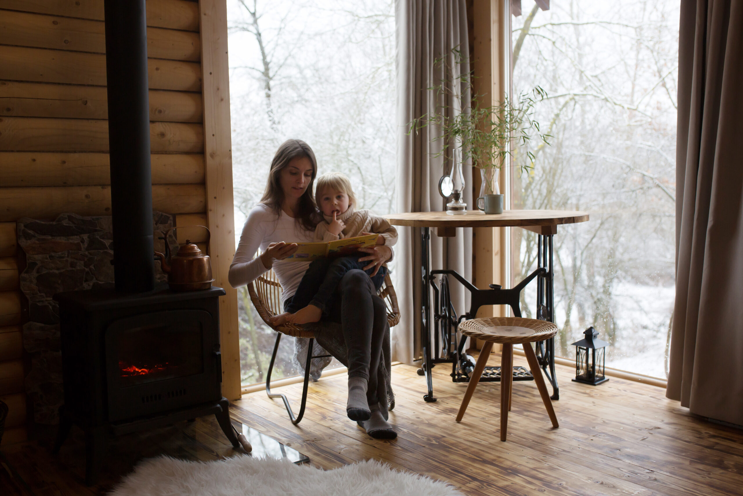 Mother and cute toddler child in a little fancy wooden cottage, reading a book, drinking tea and en...