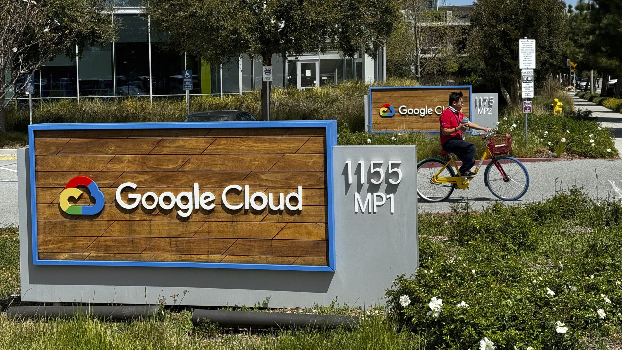 A wood sign with the Google Cloud logo on a sunny day. A person rides their bike behind it....