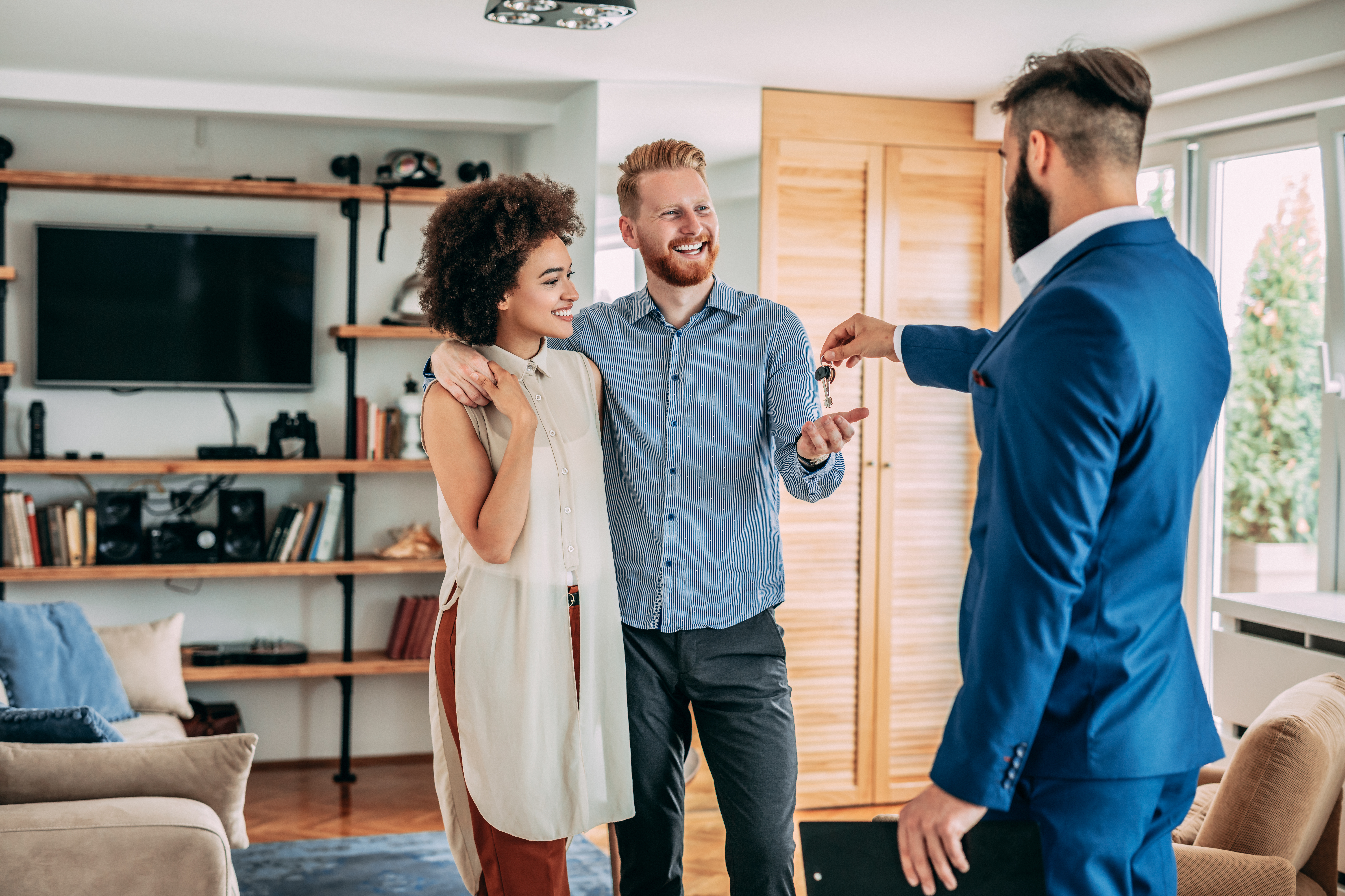 Young couple hugging while a realtor in a suit hands them keys in a new home...
