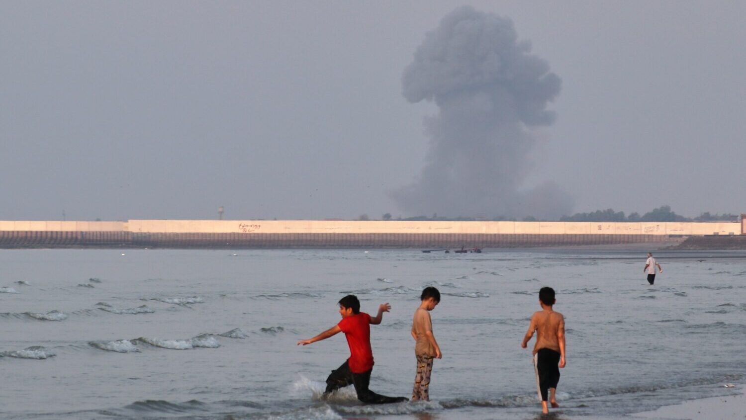 Three boys play in the shallow waters of the Strait of Hormuz, as a plume of smoke rises from an ex...